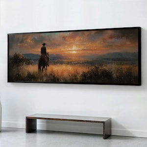 Puede incluir: Una pintura de paisaje enmarcada representa a un vaquero a caballo contra un atardecer. La obra de arte presenta tonos cálidos de naranja, amarillo y marrón, con un marco negro. La pintura se exhibe sobre un banco de madera.