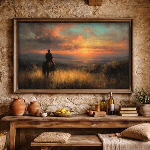 Peut inclure: Peinture encadrée d'un paysage avec un cowboy à cheval se détachant sur un coucher de soleil. L'œuvre utilise des tons chauds d'orange, de jaune et de bleu, créant un ciel dramatique. Le tableau est exposé au-dessus d'une table en bois rustique.