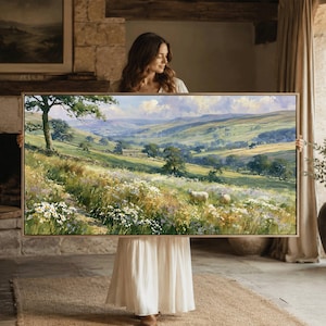 May include: A rectangular landscape painting in a light wood frame depicting a rolling green valley with a meadow of white wildflowers in the foreground, scattered trees, and two sheep grazing under a soft, cloudy sky with distant hills.