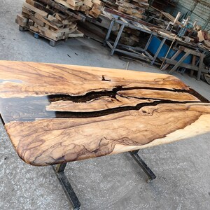 May include: A large wooden table with a natural wood grain finish. The table top has a black epoxy river running through it. The table is supported by a black metal base.