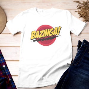 Könnte beinhalten: Weißes T-Shirt mit einer roten und gelben Grafik, auf der "BAZINGA!" steht.
