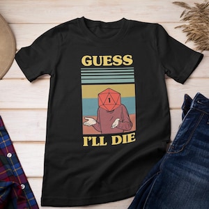 Könnte beinhalten: Schwarzes T-Shirt mit Retro-Grafik. Das Design zeigt eine Person mit einem roten Würfelkopf, der Zahl 1 und dem Text "GUESS I'LL DIE". Das Shirt wird mit Jeans, Hut und kariertem Hemd präsentiert.