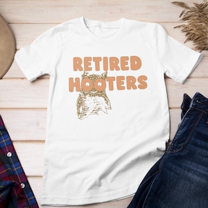 May include: White t-shirt with the text "RETIRED HOOTERS" in orange letters and an owl graphic. The shirt is displayed with blue jeans, a plaid shirt, and a brown hat on a light wood surface.