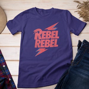 May include: Purple short-sleeved t-shirt with a red lightning bolt graphic and the text "Rebel Rebel" in red.