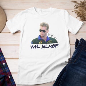 Könnte beinhalten: Weißes T-Shirt mit einem Foto von Val Kilmer, der eine Sonnenbrille und eine grüne Jacke trägt. Der Text "VAL KILMER" ist unter dem Foto aufgedruckt.