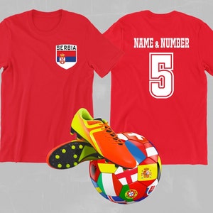 Könnte beinhalten: Rotes Fußballtrikot mit dem Wort "SERBIEN" und der serbischen Flagge auf der Vorderseite. Die Rückseite des Trikots trägt den Text "NAME & NUMMER" und die Nummer "5" in Weiß.