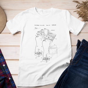 Op de afbeelding: Een wit T-shirt met een zwart-witte illustratie van een vintage videogameconsole-patent. Het patentnummer is 4,050,434. De illustratie toont de console met twee joysticks en een televisie.