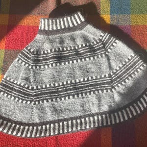 May include: A gray knitted cape with a high collar and a flared shape. The cape features horizontal bands of white and black stripes, creating a geometric pattern. The garment is displayed on a colorful plaid surface.