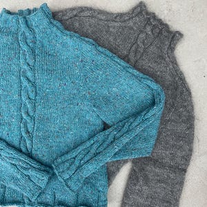 May include: Two long-sleeve jumpers with mock neck collars and cable knit detailing. One jumper is a vibrant teal blue with multicoloured flecks throughout the knit, while the second jumper is a solid charcoal grey with a textured, heathered appearance.