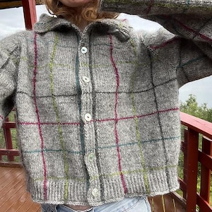 May include: A grey, button-down cardigan with a plaid pattern in red, green, and blue. The sweater has a collared neckline and is made of a thick, knitted material. The buttons are white. The cardigan is a cosy, casual garment.