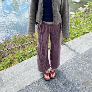 Könnte beinhalten: Eine Person trägt eine graue Strickjacke über einem marineblauen Oberteil und einer gestreiften lila Häkelhose. Rote Flip-Flops vervollständigen das Outfit. Die Person steht auf einer Betonfläche in der Nähe eines Gewässers.