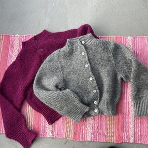 May include: Two knitted cardigans. One is a deep magenta colour, and the other is grey. Both have a high neckline and button fastenings. They are lying on a pink and white striped rug.