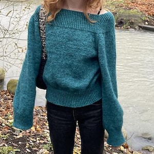 Op de afbeelding: Teal off-the-shoulder trui met lange mouwen. De trui heeft een geribbelde halslijn en manchetten. De persoon draagt een zwarte jeans. De achtergrond omvat een waterpartij en bomen.