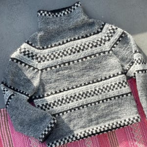 May include: A grey turtleneck jumper featuring a geometric black and white pattern. The long-sleeved jumper has a ribbed collar and is displayed on a pink and red striped textile.