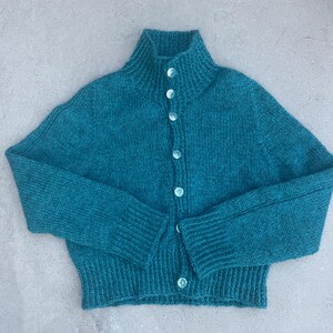 Könnte beinhalten: Eine langärmelige Strickjacke in Petrol mit hohem Stehkragen und einer durchgehenden Knopfleiste mit sechs runden, hellen Knöpfen. Das Kleidungsstück ist mit einem gerippten Saum sowie gerippten Ärmelbündchen versehen und zeigt ein Strickmuster.