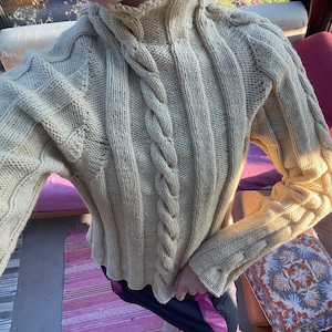 Può includere: Un maglione a maniche lunghe giallo pallido con collo alto e un evidente motivo a trecce verticali che corre lungo il centro e le maniche. Il capo è realizzato in un tessuto a maglia spesso e strutturato.