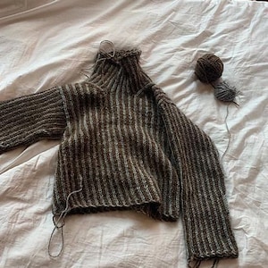 May include: A hand-knitted, brown and gray striped sweater with a turtleneck collar and long sleeves. Two balls of yarn, one brown and one gray, sit nearby. The sweater is unfinished, with loose yarn strands.
