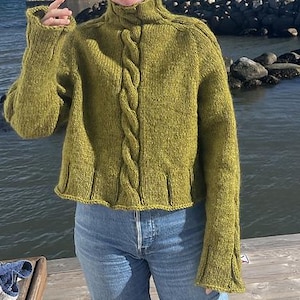 Puede incluir: Un suéter verde tejido con cuello alto y detalles de punto trenzado en la parte delantera. El suéter tiene mangas largas con puños acampanados y un dobladillo acanalado. La persona lleva vaqueros azules.