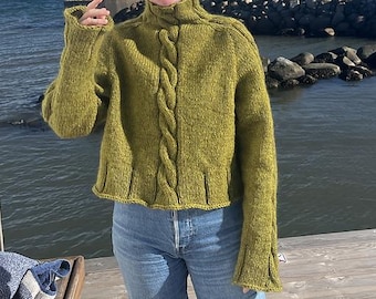 May include: A green knitted sweater with a high neck and cable knit detailing down the front. The sweater has long sleeves with flared cuffs and a ribbed hem. The person is wearing blue jeans.