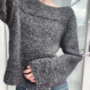 May include: A charcoal gray sweater with a wide, off-the-shoulder neckline and long sleeves. The knitwear has a textured appearance, with a mix of dark and light gray yarns. The sleeves have a ribbed cuff detail. The sweater is paired with blue jeans.