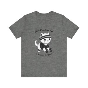 Puede incluir: Camiseta gris con un gato de dibujos animados con una cinta en la cabeza y lágrimas, levantando pesas. El texto en la camiseta dice: "Now I'm down bad crying at the gym" (Ahora estoy mal, llorando en el gimnasio).