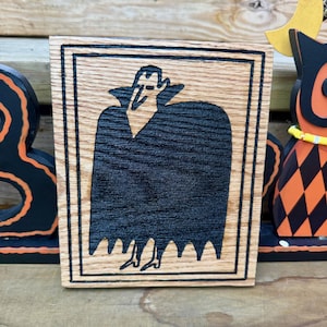 Peut inclure: Une plaque rectangulaire en bois présentant une illustration noire gravée au laser d'un vampire portant une cape à col haut. Le motif est encadré par une bordure à double ligne, mettant en valeur le grain naturel du bois clair.