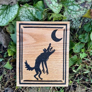 Peut inclure: Une plaque rectangulaire en bois présentant une silhouette noire gravée au laser d'un loup hurlant à la lune croissante. Le motif est entouré d'une bordure rectangulaire à double ligne, mettant en valeur le grain du bois clair.