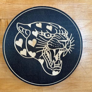 Peut inclure: Plaque ronde en bois noir avec une image grav&eacute;e d'un jaguar rugissant. Le design du jaguar int&egrave;gre des c&oelig;urs dans sa fourrure. La plaque a une bordure claire et est pos&eacute;e sur un fond en bois.