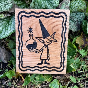 Peut inclure: Une plaque rectangulaire en bois présentant une illustration pyrogravée noire d'une créature au chapeau pointu tenant une petite bouteille avec une étoile au-dessus, le tout entouré d'une bordure décorative ondulée noire.