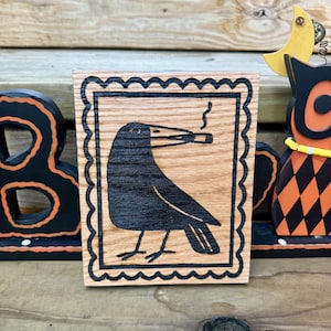 Peut inclure: Une plaque rectangulaire en bois présentant une illustration pyrogravée d'un corbeau noir tenant une cigarette allumée dans son bec. Le corbeau est centré dans une bordure festonnée décorative, le tout en lignes noires sur bois naturel.
