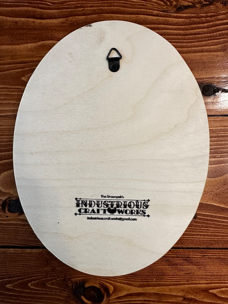 Peut inclure: Dos d'une plaque ovale en bois clair avec un crochet de suspension en m&eacute;tal noir. Le texte "The Strampek's INDUSTRIOUS CRAFT WORKS industrious.craft.works@gmail.com" est imprim&eacute; en noir.