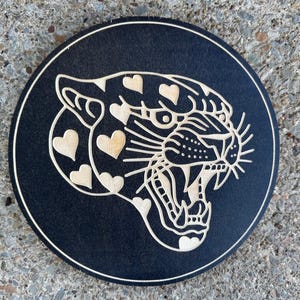 Peut inclure: Un dessous de verre rond en bois noir avec un motif d&eacute;coup&eacute; au laser d'une t&ecirc;te de jaguar rugissant. Le jaguar a des marques en forme de c&oelig;ur et est encadr&eacute; d'une fine bordure de couleur claire. Pour la d&eacute;coration int&eacute;rieure.
