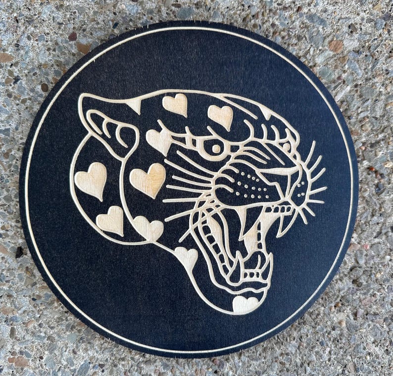 Peut inclure: Un dessous de verre rond en bois noir avec un motif d&eacute;coup&eacute; au laser d'une t&ecirc;te de jaguar rugissant. Le jaguar a des marques en forme de c&oelig;ur et est encadr&eacute; d'une fine bordure de couleur claire. Pour la d&eacute;coration int&eacute;rieure.