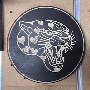 Peut inclure: Plaque ronde en bois noir avec une gravure d&eacute;taill&eacute;e d'un jaguar rugissant. La fourrure du jaguar est orn&eacute;e de motifs en forme de c&oelig;ur. Le dessin est sculpt&eacute; dans un bois clair, cr&eacute;ant un contraste saisissant.