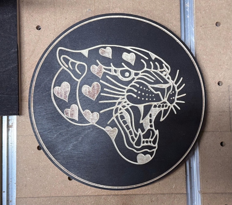 Peut inclure: Plaque ronde en bois noir avec une gravure d&eacute;taill&eacute;e d'un jaguar rugissant. La fourrure du jaguar est orn&eacute;e de motifs en forme de c&oelig;ur. Le dessin est sculpt&eacute; dans un bois clair, cr&eacute;ant un contraste saisissant.