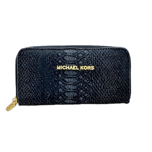 May include: Black zip-around wallet with a snakeskin texture. The wallet features the gold text "MICHAEL KORS" on the front. A gold zipper pull is visible on the left side. The wallet is rectangular and appears to be made of leather.