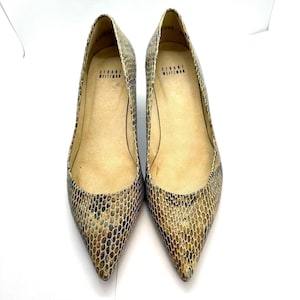 May include: A pair of Stuart Weitzman pointed-toe pumps. The shoes have a snakeskin pattern in brown, tan, and black. The interior of the shoes is light tan. The brand name is visible inside the shoe.