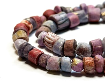 May include: A strand of colorful, cylindrical beads in shades of red, purple, blue, and brown. Some beads have gold accents. The beads are strung together, creating a curved shape against a white background. The beads appear to be made of stone or glass.