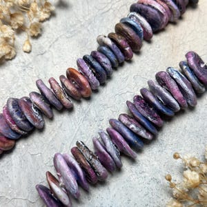 May include: Two strands of colourful, flat, oval-shaped beads in shades of purple, blue, and brown. The beads have a natural, organic appearance, with variations in colour and texture. The beads are arranged in a row, suggesting they are for jewellery making.