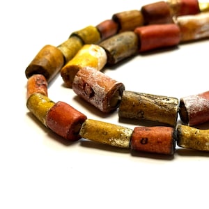 May include: A strand of colourful, cylindrical beads made of natural materials. The beads are a mix of yellow, orange, and brown, and have a rustic, aged appearance.
