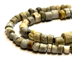 May include: A strand of antique bone beads in shades of grey, brown, and white. The beads are irregular in shape and size, giving the strand a rustic and unique look.