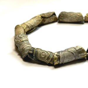 May include: A close-up of a necklace made of cylindrical beads with a gold-like finish. The beads are decorated with intricate patterns and swirls.