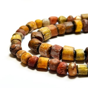 May include: A necklace of cylindrical beads in various earth tones, including shades of brown, orange, yellow, and red. The beads have a textured, weathered appearance, suggesting they are made of natural materials. The beads are strung together, forming a curved line against a white background.