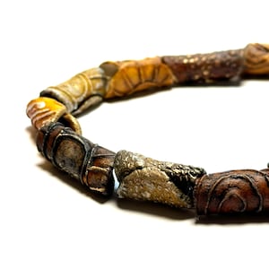 May include: A close-up of a necklace with cylindrical beads in shades of brown, tan, and gold. The beads have a textured, possibly carved, appearance. The necklace is set against a white background.