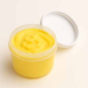 Könnte beinhalten: Ein durchsichtiges Plastikgefäß gefüllt mit einer leuchtend gelben, cremigen Substanz. Der weiße Deckel ist leicht geöffnet und zeigt die Textur des Produkts. Es handelt sich wahrscheinlich um eine Hautcreme oder ein Kosmetikprodukt.