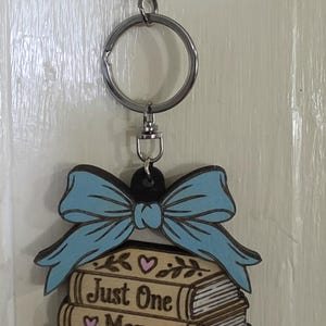 May include: A keychain featuring a stack of three books with a light blue bow on top. The books are engraved with the text "Just One More Chapter" and decorated with small pink hearts and leaf illustrations. The keychain has a silver metal ring and clasp.