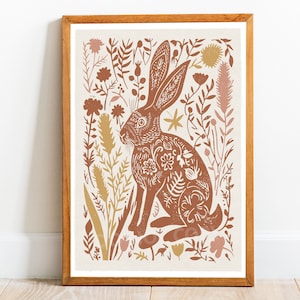 May include: A framed art print featuring a brown rabbit with floral patterns, surrounded by stylized plants and flowers in shades of brown, tan, and pink. The artwork is set against a cream background, framed in a wooden frame.