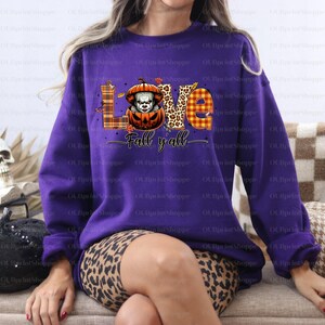 May include: A purple sweatshirt featuring a Halloween design. The graphic spells "LOVE" with a pumpkin-clown in place of the 'O'. The words "Fall y'all" are written below. The design incorporates autumnal colours and plaid patterns.