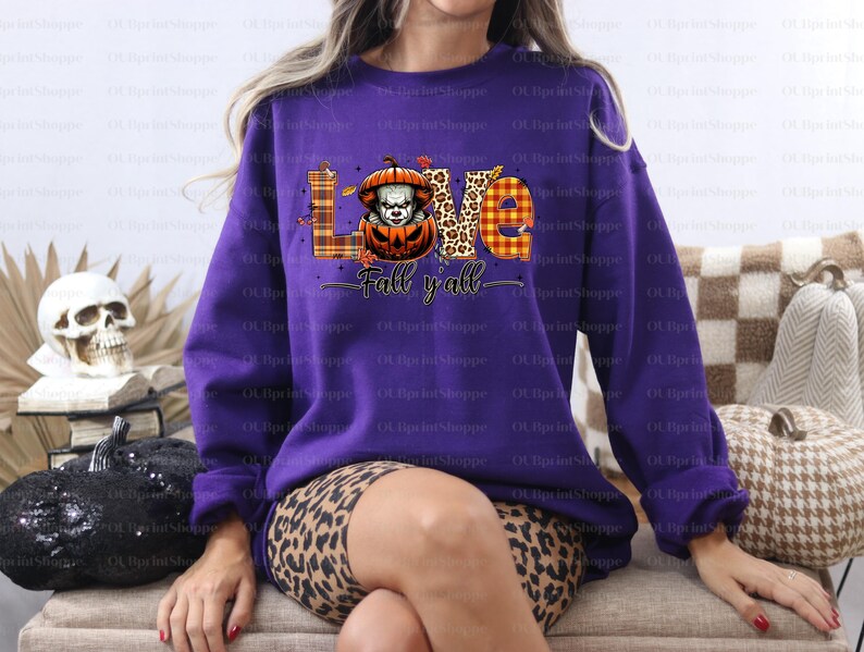 May include: A purple sweatshirt featuring a Halloween design. The graphic spells "LOVE" with a pumpkin-clown in place of the 'O'. The words "Fall y'all" are written below. The design incorporates autumnal colours and plaid patterns.
