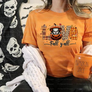 May include: Orange t-shirt with the word "LOVE" in a mix of patterns and a pumpkin with a scary clown face. The text "Fall y'all" is below the word "LOVE". A matching orange mug with "PUMPKIN SPICE" is held in the hand.